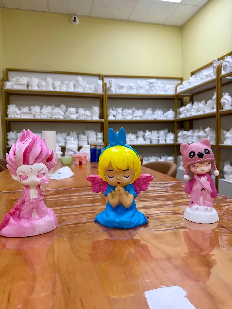 Three hand-painted figures in pink, blue and yellow with plaster blanks on shelves behind them at Craft Pop